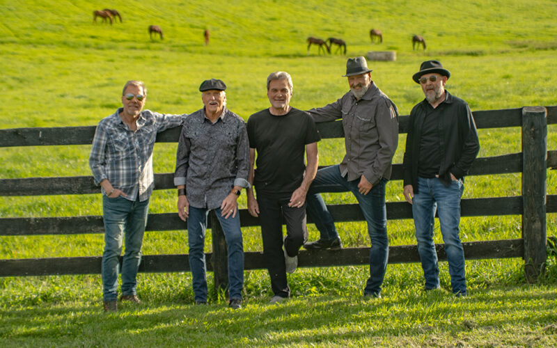The five members of Exile standing beside a wooden fence in a green pasture with horses.