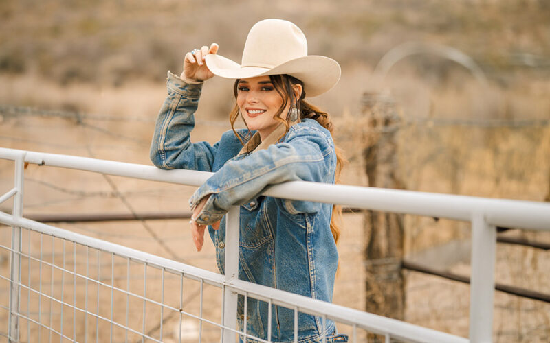 Jenna Paulette in a white cowboy hat and denim jacket, leaning on a ranch gate.
